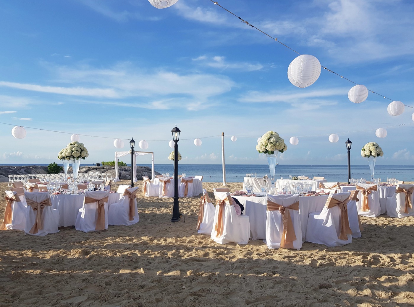Beach wedding reception beside the ocean with white lanterns and gold chair ribbons